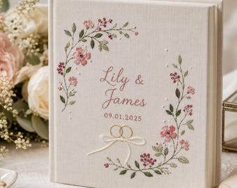 May include: A square, cream-colored fabric-covered photo album featuring embroidered floral vines in shades of pink and green. The cover displays the text Lily & James, the date 09.01.2025, and an embroidered design of two interlocking rings.