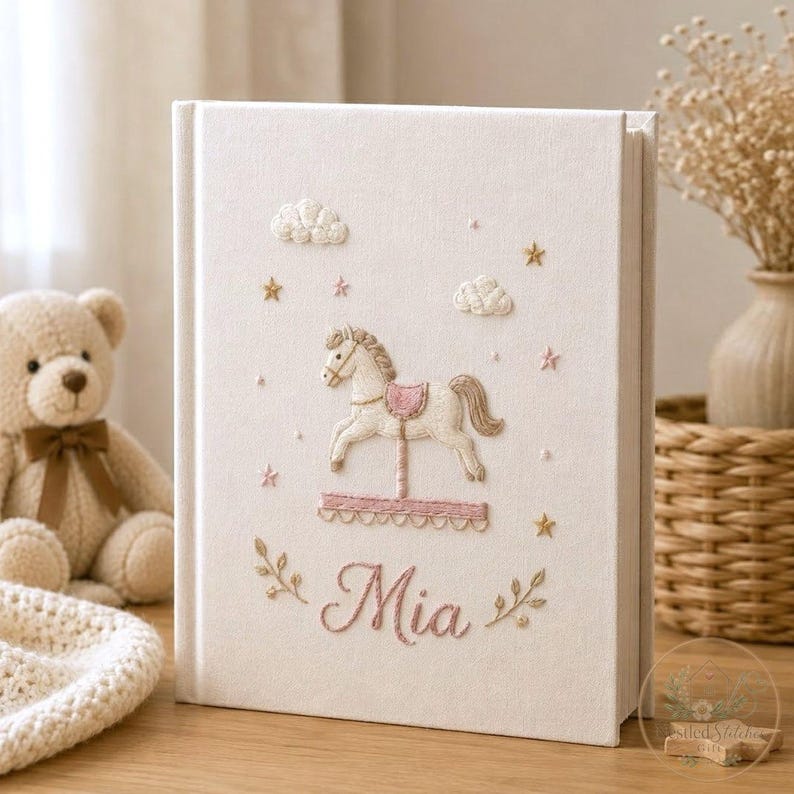 May include: A cream-colored fabric-covered book featuring embroidered details on the front cover, including a carousel horse, clouds, stars, and the name Mia in pink script, accented by small leafy branches at the bottom of the cover design.