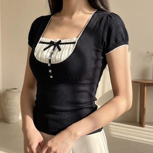 Op de afbeelding: Een zwart gebreid shirt met korte mouwen en een vierkante halslijn met witte rand. Het middenpaneel is gemaakt van witte geplooide stof, versierd met een kleine zwarte strik aan de bovenkant en een verticale rij van vier ronde, fonkelende knopen.