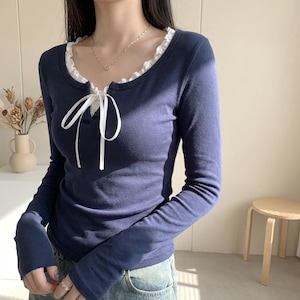 May include: A long-sleeved navy blue top featuring a scoop neckline with white lace trim and a white ribbon tie at the center. The garment has a fitted silhouette and is paired with blue denim jeans, showcasing a simple and clean design.