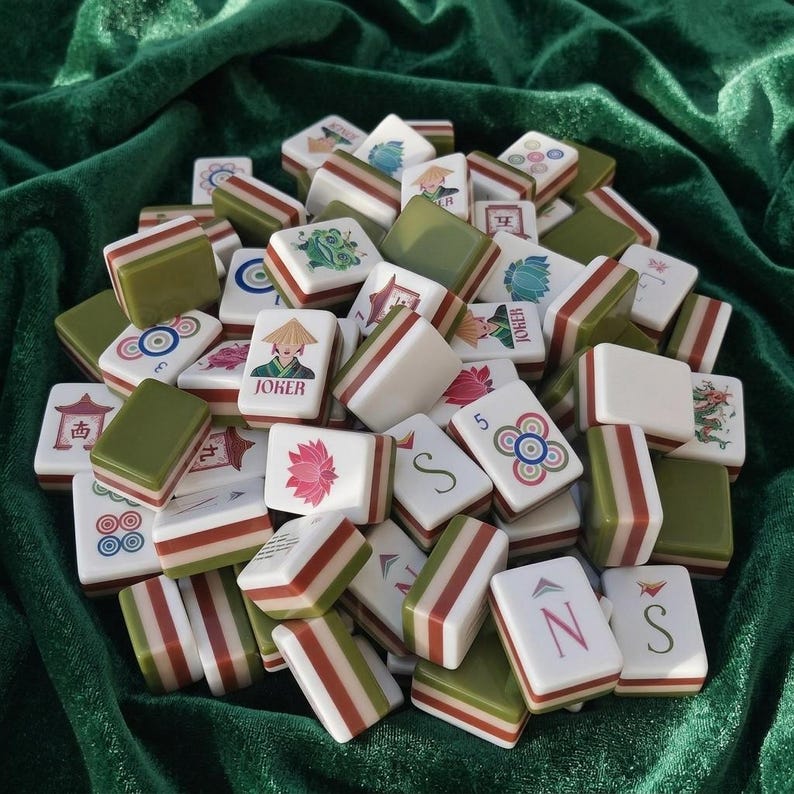 Mahjong tile set piled on green velvet fabric, featuring white and olive green resin tiles with red stripe detailing, decorated with colorful Asian-inspired illustrations including joker tiles, lotus flowers, dragons, pagodas, circles, and compass direction letters, American mahjong game set with complete tile collection