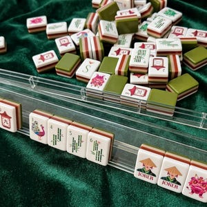 American mahjong tile set with clear acrylic racks displayed on green velvet surface, featuring colorful illustrated tiles with joker, peacock, dragon and bamboo suit designs, cream white resin tiles with olive green and red stripe backs, complete mah jongg game set ready to play