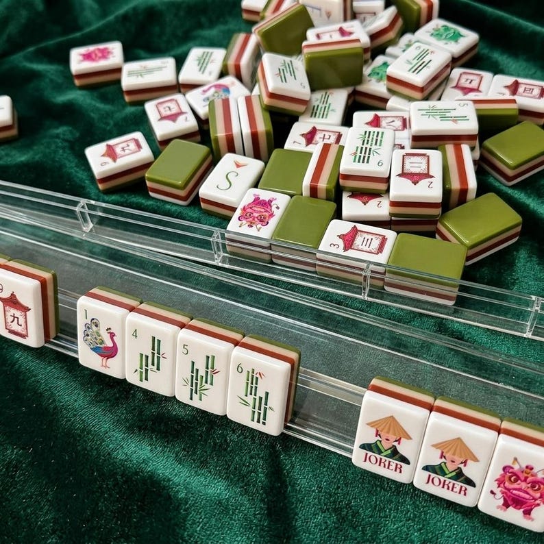 American mahjong tile set with clear acrylic racks displayed on green velvet surface, featuring colorful illustrated tiles with joker, peacock, dragon and bamboo suit designs, cream white resin tiles with olive green and red stripe backs, complete mah jongg game set ready to play
