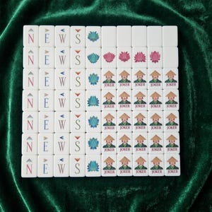 American Mahjong tile set displayed on green velvet showing white tiles with NEWS wind tiles, lotus flower tiles in teal and pink, and joker tiles featuring an Asian figure in a conical hat, arranged in a full grid layout for easy viewing of complete tile designs in a modern decorative style