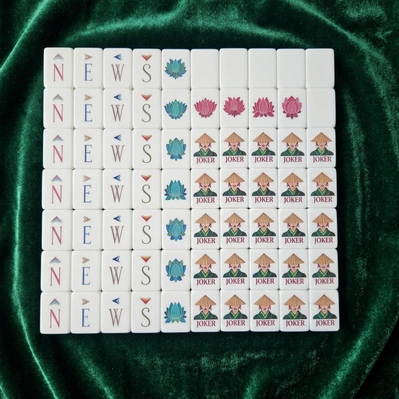 American Mahjong tile set displayed on green velvet showing white tiles with NEWS wind tiles, lotus flower tiles in teal and pink, and joker tiles featuring an Asian figure in a conical hat, arranged in a full grid layout for easy viewing of complete tile designs in a modern decorative style