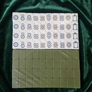 Mahjong tile set displayed on green velvet fabric showing white tiles with colorful circle suit symbols numbered 1 to 9 arranged in four rows alongside olive green back-facing tiles, complete traditional Chinese mahjong game set with numbered dot tiles and pagoda design, suitable for family game nights and tournaments
