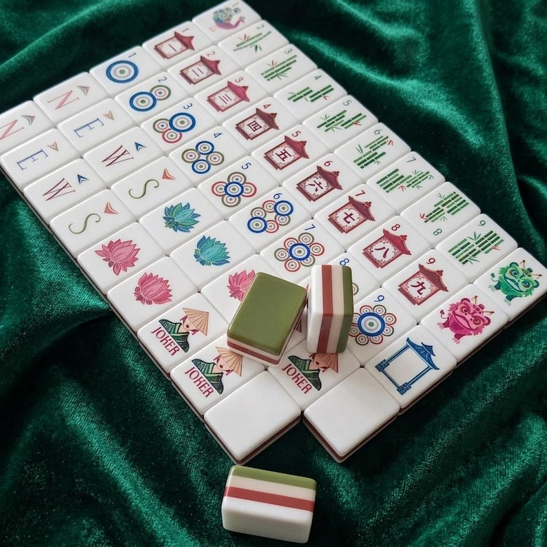 Mahjong tile set displayed on green velvet fabric featuring white tiles with colorful illustrations including lotus flowers, bamboo, circles, Chinese characters, joker tiles, and wind suit letters, accompanied by two-tone green and pink striped dice, complete American mahjong set with decorative floral and Asian-inspired artwork