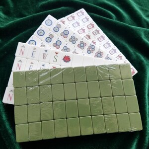 Mahjong tile set with white engraved tiles featuring circle suit, bamboo suit, and wind tiles with colorful lotus flower design, displayed alongside olive green blank back tiles wrapped in plastic, arranged on dark green velvet fabric, complete Chinese mahjong game set for traditional tile-based strategy gameplay