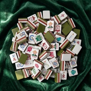 Mahjong tile set with colorful illustrated designs including joker tiles, lotus flowers, dragon, and Chinese characters, featuring olive green and terracotta striped backs, piled on green velvet fabric, complete American mahjong set with decorative artwork style tiles for game night or gift giving