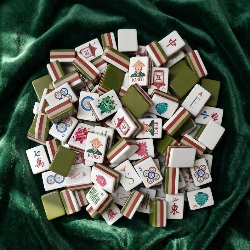 Mahjong tile set with colorful illustrated designs including joker tiles, lotus flowers, dragon, and Chinese characters, featuring olive green and terracotta striped backs, piled on green velvet fabric, complete American mahjong set with decorative artwork style tiles for game night or gift giving
