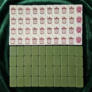 Mahjong tiles set displayed on green velvet surface showing cream colored numbered character tiles 1 through 9 with Chinese pagoda and lion dance designs arranged in four rows alongside olive green blank tile backs, complete mahjong game set with traditional Asian artwork suitable for family board game nights