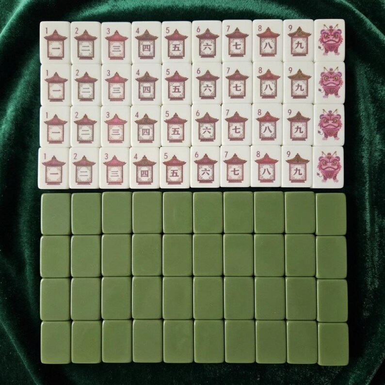 Mahjong tiles set displayed on green velvet surface showing cream colored numbered character tiles 1 through 9 with Chinese pagoda and lion dance designs arranged in four rows alongside olive green blank tile backs, complete mahjong game set with traditional Asian artwork suitable for family board game nights
