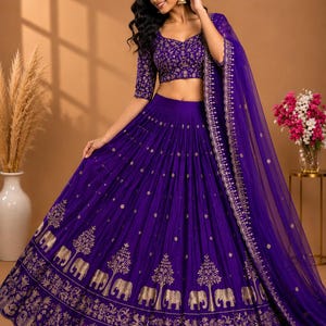 May include: A deep purple lehenga choli set featuring a cropped blouse with short sleeves and a voluminous floor-length skirt. Both pieces are adorned with intricate gold embroidery, including elephant motifs and tree patterns along the hem.