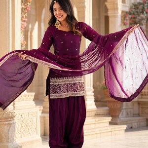 May include: A deep purple salwar kameez set featuring a long-sleeved tunic with a square neckline, small embroidered floral motifs, and a wide metallic border at the hem. The outfit includes matching loose-fitting trousers and a sheer dupatta.