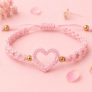 May include: A light pink woven cord bracelet featuring a central heart-shaped charm made of matching pink thread. The bracelet is accented with small, polished gold-tone metal beads integrated into the woven band and has an adjustable sliding knot closure.