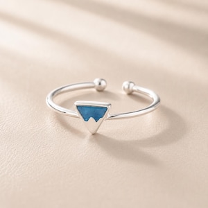 May include: A silver open-band ring featuring a triangular centrepiece with a blue mountain-shaped inlay. The band is thin and polished, ending in two small spherical terminals at the opening. The design is simple, metallic, and reflective.