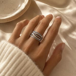 Puede incluir: Un anillo de plata con un diseño cruzado de múltiples hilos. La banda consta de varios alambres metálicos entrelazados, con algunos hilos que muestran un acabado pulido liso y otros que presentan un detalle de superficie delicadamente perlado o texturizado.
