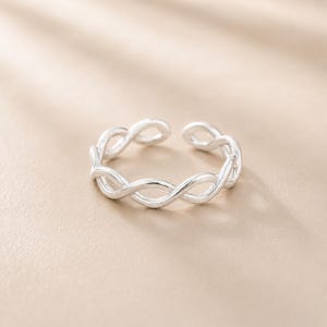 May include: A silver-toned ring featuring an open, adjustable band design with a continuous twisted, rope-like pattern. The metal surface is smooth and reflective, forming a circular shape with a small gap at the back for size adjustment.