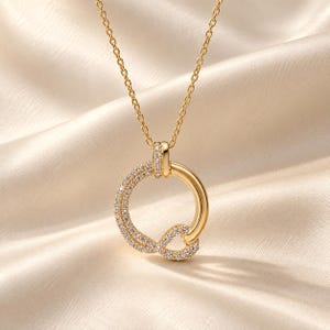 Puede incluir: Un collar de tono dorado con un colgante circular de diseño trenzado. Una mitad del círculo es de oro pulido liso, mientras que la otra mitad está incrustada con pequeñas piedras claras y brillantes. El colgante cuelga de una delicada cadena dorada.