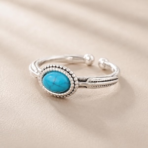 Peut inclure: Une bague ajustable en argent ornée d'une pierre turquoise ovale avec des veines sombres. La pierre est sertie dans une bordure perlée, et l'anneau présente un motif texturé en forme de plume de chaque côté du sertissage central.