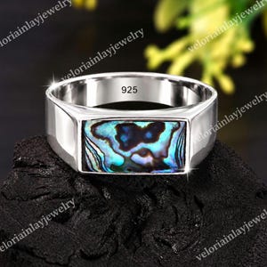 May include: A silver ring featuring a rectangular inlay of iridescent abalone shell with swirling blue, green, and black patterns. The interior of the band is clearly stamped with the text 925, indicating sterling silver composition.