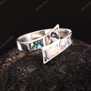 Op de afbeelding: Een zilveren ring met een geometrisch ontwerp met drie ingelegde iriserende abalone schelpsegmenten. De metalen band heeft een gepolijste afwerking en de schelpstukjes vertonen verschuivende tinten blauw, groen en paars in het zilver.