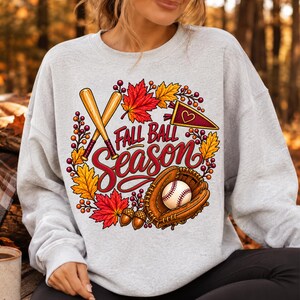 Op de afbeelding: Een lichtgrijs sweatshirt met ronde hals en een grafische print met de tekst Fall Ball Season. Het ontwerp bevat twee gekruiste houten honkbalknuppels, een honkbal in een leren handschoen, herfstbladeren, eikels en een klein wimpeltje.