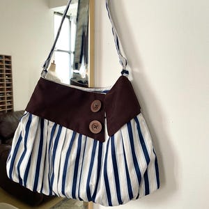 May include: A hobo-style shoulder bag featuring a white and navy blue vertical striped pattern. The bag has a wide, solid dark brown fabric band across the top front, accented with two large, round, brown buttons. It includes a matching striped shoulder strap.