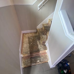 May include: A staircase with exposed wooden treads showing remnants of old adhesive and padding. The stairs are flanked by light beige walls with white trim and a white handrail. The steps appear to be in a state of renovation or repair.
