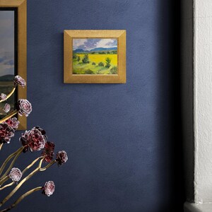 Original New Mexico landscape oil painting Abiquiu Field of Summer Gold by Dawn Chandler hanging in a gold frame on a deep blue wall, paired with sculptural purple succulents in the foreground and a glimpse of another landscape, demonstrating how the glowing yellow field and green trees enliven contemporary Southwestern décor.