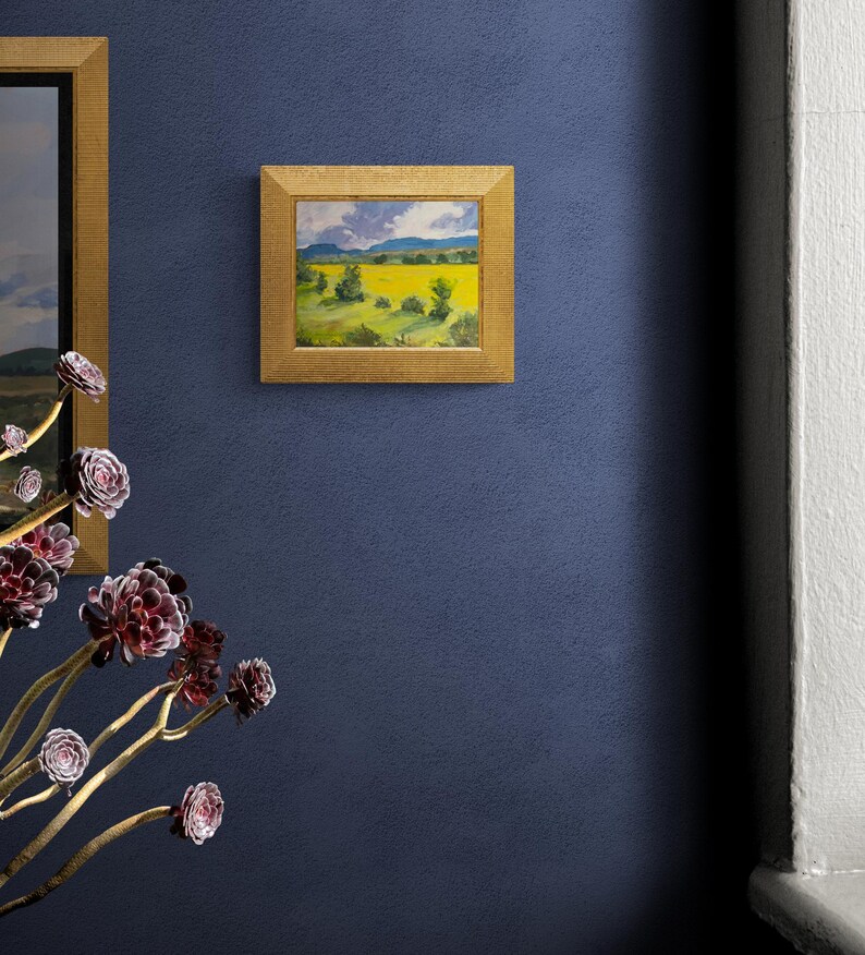 Original New Mexico landscape oil painting Abiquiu Field of Summer Gold by Dawn Chandler hanging in a gold frame on a deep blue wall, paired with sculptural purple succulents in the foreground and a glimpse of another landscape, demonstrating how the glowing yellow field and green trees enliven contemporary Southwestern décor.