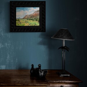 Original New Mexico landscape oil painting “Finding My Way to Pedernal” by Santa Fe artist Dawn Chandler, shown framed in an ornate black frame above a rustic wooden dresser with sculptural black bird figures and a dark table lamp, illustrating how this vibrant Southwest mesa and valley scene enriches a cozy, moody interior.