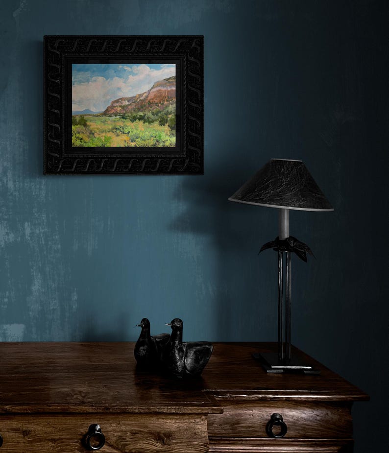 Original New Mexico landscape oil painting “Finding My Way to Pedernal” by Santa Fe artist Dawn Chandler, shown framed in an ornate black frame above a rustic wooden dresser with sculptural black bird figures and a dark table lamp, illustrating how this vibrant Southwest mesa and valley scene enriches a cozy, moody interior.