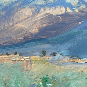 Original New Mexico landscape oil painting Spring Morning Over the Taos Mountains by Santa Fe artist Dawn Chandler shown framed in a warm golden frame and hanging in a recessed blue-gray niche above three simple wooden vessels, illustrating how the colorful Taos valley, blue mountains, and dramatic sky bring vibrant Southwestern mountain art and a sense of spacious calm to a contemporary interior wall.