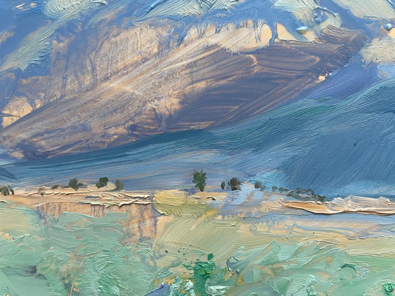 Original New Mexico landscape oil painting Spring Morning Over the Taos Mountains by Santa Fe artist Dawn Chandler shown framed in a warm golden frame and hanging in a recessed blue-gray niche above three simple wooden vessels, illustrating how the colorful Taos valley, blue mountains, and dramatic sky bring vibrant Southwestern mountain art and a sense of spacious calm to a contemporary interior wall.