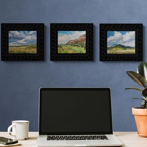 Trio of original New Mexico landscape oil paintings by Santa Fe artist Dawn Chandler, featuring “Finding My Way to Pedernal” centered between two companion Southwest sky and mesa scenes, all in carved black frames above a minimalist desk with laptop, mug and potted rubber plant, suggesting how this art elevates a modern home office.