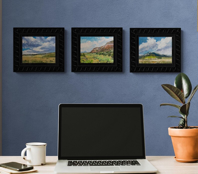 Trio of original New Mexico landscape oil paintings by Santa Fe artist Dawn Chandler, featuring “Finding My Way to Pedernal” centered between two companion Southwest sky and mesa scenes, all in carved black frames above a minimalist desk with laptop, mug and potted rubber plant, suggesting how this art elevates a modern home office.