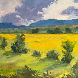 Abiquiu Field of Summer Gold, an original New Mexico landscape oil painting by Santa Fe artist Dawn Chandler, displayed in a black ornate frame on a plum-colored wall above a rustic wooden table with books, small ceramic vases, and a bowl of seed pods, illustrating how the sunny field scene warms an intimate reading nook.