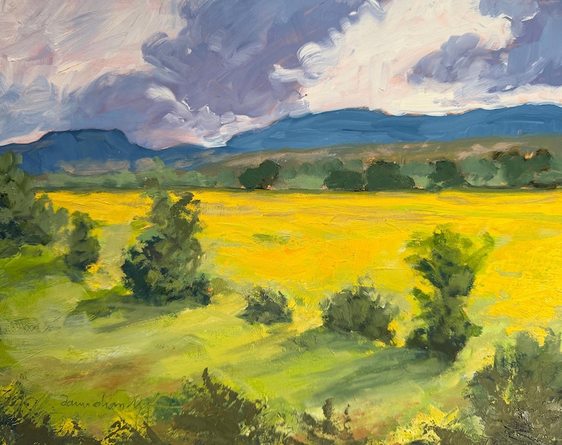 Abiquiu Field of Summer Gold, an original New Mexico landscape oil painting by Santa Fe artist Dawn Chandler, displayed in a black ornate frame on a plum-colored wall above a rustic wooden table with books, small ceramic vases, and a bowl of seed pods, illustrating how the sunny field scene warms an intimate reading nook.