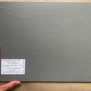 Back of original New Mexico landscape oil painting Spring Morning Over the Taos Mountains by Santa Fe artist Dawn Chandler, showing the gray-primed 9 x 12 inch panel held at the corner by the artist’s hand, with a neatly labeled sticker indicating title, medium, size, and Dawn Chandler’s Santa Fe contact information, emphasizing the authenticity and professional presentation of the artwork.