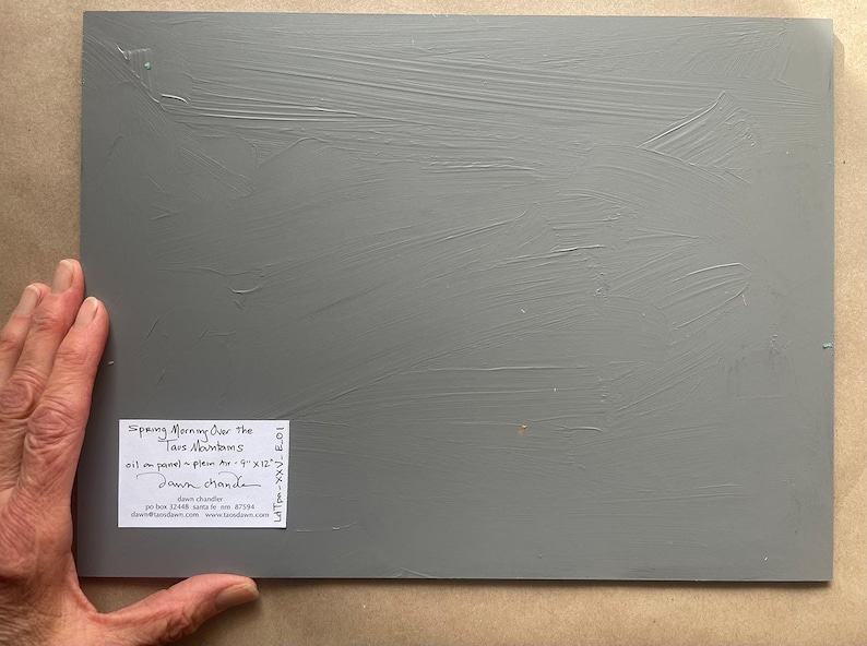 Back of original New Mexico landscape oil painting Spring Morning Over the Taos Mountains by Santa Fe artist Dawn Chandler, showing the gray-primed 9 x 12 inch panel held at the corner by the artist’s hand, with a neatly labeled sticker indicating title, medium, size, and Dawn Chandler’s Santa Fe contact information, emphasizing the authenticity and professional presentation of the artwork.
