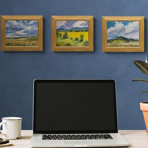 Three framed original New Mexico landscape oil paintings by Santa Fe artist Dawn Chandler, including Abiquiu Field of Summer Gold in the center, hanging on a blue wall above a minimalist desk with laptop, mug, phone, and potted rubber plant, suggesting how these small Southwest artworks brighten a home office or studio.