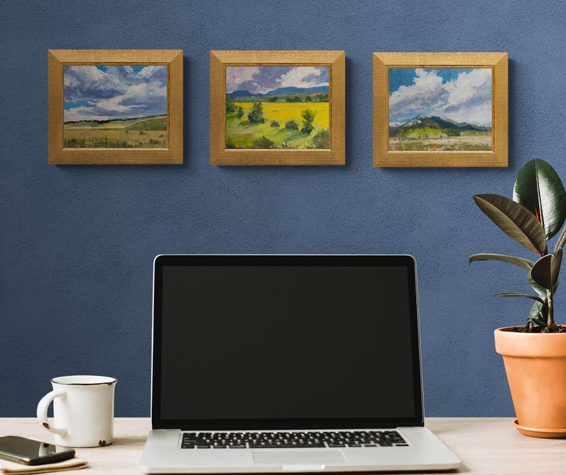 Three framed original New Mexico landscape oil paintings by Santa Fe artist Dawn Chandler, including Abiquiu Field of Summer Gold in the center, hanging on a blue wall above a minimalist desk with laptop, mug, phone, and potted rubber plant, suggesting how these small Southwest artworks brighten a home office or studio.