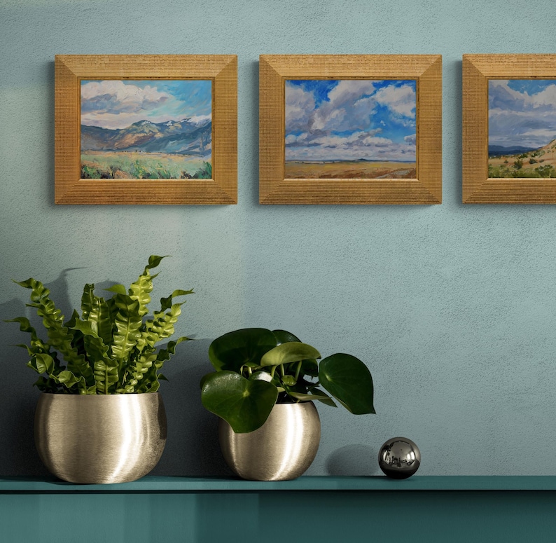 Stylish interior view with three original New Mexico landscape paintings by Santa Fe artist Dawn Chandler, including Spring Morning Over the Taos Mountains, all framed in warm golden frames and hung on a soft blue wall above metallic planters filled with lush green houseplants, showing how the expressive mountain skies and high-desert fields complement modern Southwestern decor and add a fresh, nature-inspired focal point.
