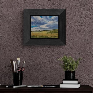 Room view of Santa Fe artist Dawn Chandler’s original New Mexico landscape oil painting Under a High Country August Sky, shown framed in a dark, wide wood frame and hanging on a textured plum‑colored wall above a desk with brushes, drawing tools, books, and a potted plant, demonstrating how her New Mexico landscape paintings bring expansive sky and meadow vistas into a contemporary home or studio setting.