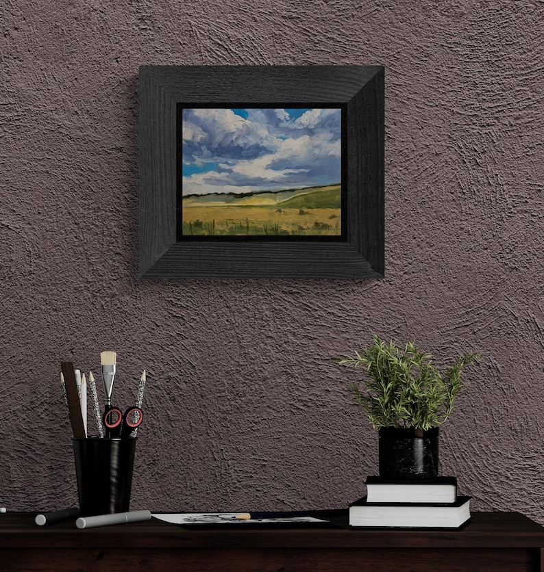 Room view of Santa Fe artist Dawn Chandler’s original New Mexico landscape oil painting Under a High Country August Sky, shown framed in a dark, wide wood frame and hanging on a textured plum‑colored wall above a desk with brushes, drawing tools, books, and a potted plant, demonstrating how her New Mexico landscape paintings bring expansive sky and meadow vistas into a contemporary home or studio setting.