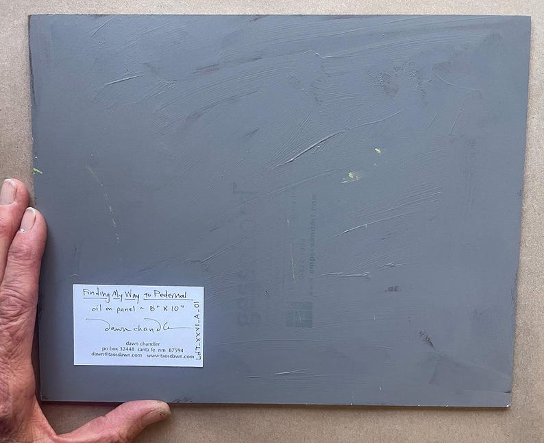 Back of original New Mexico landscape oil painting “Finding My Way to Pedernal” by Santa Fe artist Dawn Chandler, showing the gray-painted 8 x 10 inch panel held at the lower left by the artist’s hand, with a white signed label identifying the title, medium, size and Dawn Chandler’s Santa Fe contact information, confirming this is a one-of-a-kind artwork.