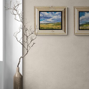 Stylish neutral interior with a tall ceramic vase holding a delicate bare branch on a ledge beside two framed original New Mexico landscape paintings by Santa Fe artist Dawn Chandler, including Under a High Country August Sky in a light wood frame with black inner liner, the artwork’s sweeping clouds and golden fields bringing movement and color to the soft beige wall while coordinating with another New Mexico sky and mountain painting.