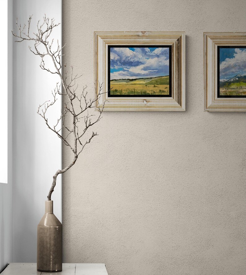 Stylish neutral interior with a tall ceramic vase holding a delicate bare branch on a ledge beside two framed original New Mexico landscape paintings by Santa Fe artist Dawn Chandler, including Under a High Country August Sky in a light wood frame with black inner liner, the artwork’s sweeping clouds and golden fields bringing movement and color to the soft beige wall while coordinating with another New Mexico sky and mountain painting.
