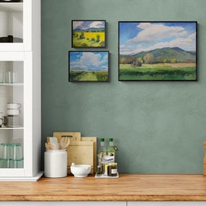 Original New Mexico landscape oil paintings by Dawn Chandler, including Abiquiu Field of Summer Gold, arranged as a small gallery wall in a sage green kitchen above a wooden countertop with cutting boards, glassware, and olive oil bottles, showing how her bright Southwest skies and fields add color to everyday spaces.